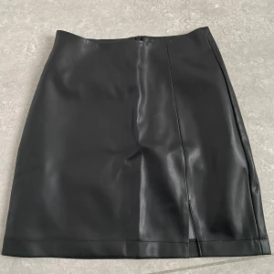 Svart kjol från BikBok - Snygg svart kjol i skinnimitation från BikBok. Kjolen har en diskret slits framtill och dragkedja baktill. Endast använd en gång. Nyskick.