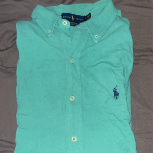 Turkos skjorta från Ralph Lauren - Säljer en snygg turkos skjorta från Ralph Lauren med klassisk krage och knappar framtill. Skjortan har en broderad logga i mörkblått på bröstet och är tillverkad i ett lätt och luftigt material. Passar (M&S )