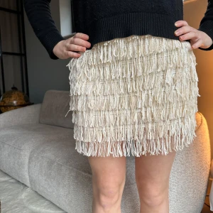 Minikjol med fransar - Snygg beige kortkjol täckt med glittrande paljetter och fransar. Perfekt för att sticka ut med en unik look. Kjolen har en tight passform och är idealisk för festliga tillfällen.