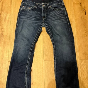 Mörkblå jeans från True Religion - Snygga mörkblå jeans från True Religion med kontrastsömmar och broderade bakfickor. Jeansen har en klassisk femficksdesign och en knappgylf. Perfekta för en avslappnad stil.