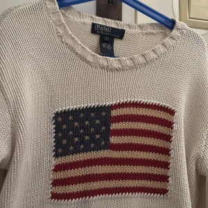 Vit stickad tröja från Polo Ralph Lauren - Säljer en vit stickad tröja från Polo Ralph Lauren med ett stort amerikanskt flaggmotiv på framsidan. Tröjan har långa ärmar och en rund halsringning. Perfekt för en stilren och avslappnad look. 🌟Denna är i barn storlek btw pris kan diskuteras eftersom den har en blå liten fläck på framsidan som man nästan dock inte kan se!