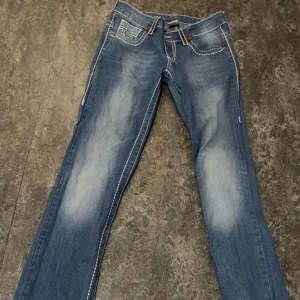 Blå jeans från True Religion - Snygga blå jeans från True Religion med distinkta vita sömmar och broderade detaljer på bakfickorna. De har en klassisk femficksdesign och en bootcut/straight-stil som ger en avslappnad look. Perfekta för en casual outfit. Dock så finns det downturns, byxan har ett litet hål i backfickan men det inget stort problem, går fortfarande att fixa genom att sy. Pris kan diskuteras ! (: byxan är storlek L men kan även passa som M ! 