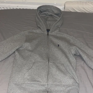 Grå hoodie från Polo Ralph Lauren - Säljer en stilren grå hoodie från Polo Ralph Lauren med dragkedja och klassisk logga på bröstet. Perfekt för en avslappnad look. Den har en bekväm passform och är tillverkad i mjukt material.