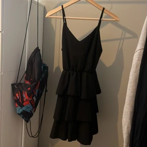 Svart klänning med volanger - Snygg svart klänning med smala axelband och v-ringning. Den har flera lager av volanger som ger en fin rörelse. Perfekt för en kväll ute eller en speciell tillställning.