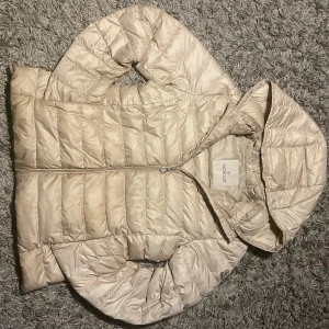 Beige dunjacka från Moncler - Säljer en snygg beige dunjacka från Moncler med huva och dragkedja. Jackan har en quiltad design som ger en stilren look. Perfekt för kyligare dagar.