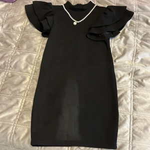 Svart klänning med volangärmar och pärldetalj - Elegant svart klänning med volangärmar och en fin pärldetalj vid halsen. Klänningen har en dragkedja i ryggen för enkel påklädning. Perfekt för en stilren look.