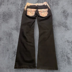 Miss Me Byxor med blommigt motiv  - Unika svarta byxor från Miss Me med blommiga detaljer i rosa. Byxorna har en lågmidjad passform och bootcut-stil. Längd: 99cm Innerbenslängd: 79cm Midja: 36-40cm (strechiga)