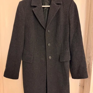 Grå kappa med knappar - Elegant grå kappa med klassisk design och knappar framtill. Den har en stilren look med långa ärmar och en rak passform. Perfekt för kyligare dagar.