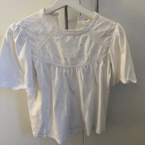 Vit blus från Zara - Säljer en söt vit blus från Zara med korta ärmar och vackra broderade detaljer på bröstet. Perfekt för en stilren och elegant look. Blusen har en rund halsringning och är gjord i ett mjukt material.