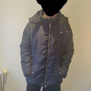 Svart dunjacka från H&M - Säljer en svart dunjacka från H&M med huva och dragkedja. Jackan har en quiltad design och långa ärmar, perfekt för kyliga dagar. Den är både stilren och funktionell.