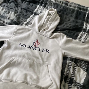 Vit hoodie från Moncler - Säljer en stilren vit hoodie från Moncler med stor logotyp på framsidan. Hoodien har en klassisk känguruficka och justerbar huva med dragsko. Perfekt för en avslappnad och trendig look.