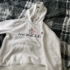Vit hoodie från Moncler - Säljer en stilren vit hoodie från Moncler med broderad logotyp på bröstet. Hoodien har en klassisk känguruficka och justerbar huva med dragsko. Perfekt för en avslappnad och trendig look. QR. Kod finns 