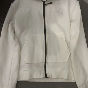  C.P. Company kofta - Säljer en stilren vit huvtröja från C.P. Company med dragkedja och broderad logga på bröstet. Tröjan har en unik detalj med glasögon i huvan och praktiska fickor framtill. Perfekt för en cool och avslappnad stil. Jag fick den som födelsedags present men jag gillar inte den så mycket och har aldrig haft på mig den ute bara testat på mig. 