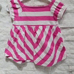 Barnklänning - Söt liten klänning i rosa och vit med ränder från Petter & Kajsa. Den har korta ärmar och en luftig passform, perfekt för varma dagar. Klänningen har en lekfull design med diagonala ränder på kjolen och horisontella ränder på toppen.