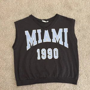 Svart ärmlös topp från KappAhl - Säljer en svart ärmlös topp från KappAhl med texten 'MIAMI 1990' i ljusblått och vitt på framsidan. Toppen har en v-ringad hals och ribbad nederkant. Perfekt för en avslappnad stil.