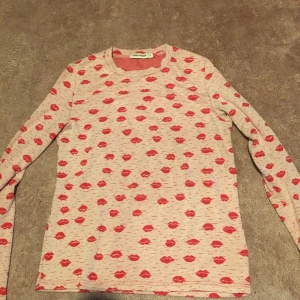 Ljusrosa tröja med röda läppar från Carin Wester - Säljer en  långärmad tröja från Carin Wester med ett coolt mönster av röda läppar. Perfekt för dig som vill sticka ut med en unik stil. Tröjan är i storlek S och har en normal passform.