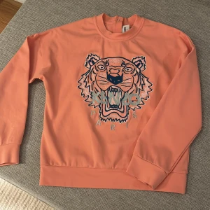 Orange sweatshirt från Kenzo Kids - Säljer en cool orange sweatshirt från Kenzo Kids med ett stort tigertryck och texten 'Kenzo Paris' på framsidan. Tröjan har långa ärmar och en rund halsringning. Perfekt för att ge en färgklick till garderoben! Har används 1 gång och är i super bra skick!! Frakt tillkommer!