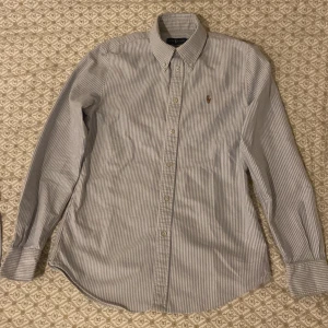 Randig skjorta från Ralph Lauren - Snygg randig skjorta från Ralph Lauren i blått och vitt. Skjortan har långa ärmar och klassisk krage med knappar. Den är broderad med den ikoniska logotypen på bröstet. Perfekt för en stilren look. Storlek S/XS. 