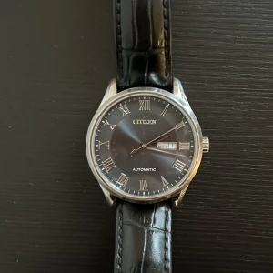Svart klocka från Citizen - Elegant klocka från Citizen med svart urtavla och romerska siffror. Den har en automatisk rörelse och datumvisning. Armbandet är i svart skinn med krokodilpräglat mönster. Perfekt för den stilmedvetna! Pris kan discuteras 