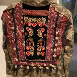 Camouflagejacka med broderade detaljer - Camouflagejacka med broderade blommor och tofsar på ryggen. Jackan har en cool kombination av militärstil och färgglada detaljer. 
