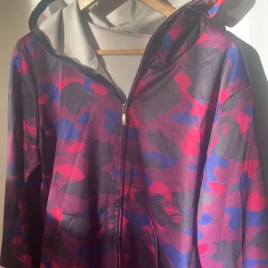 Flerfärgad bape hoodie med dragkedja (ej äkta) - Säljer en cool flerfärgad hoodie med camo-mönster i rosa, blått och svart. Hoodien har en dragkedja framtill och en unik design på huvan med tuffa detaljer. Perfekt för dig som vill sticka ut! Storlek L men passar även M