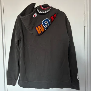 Bape Shark Hoodie - En bape shark hoodie med asfet wash, hoodien är grå/mullvad i färgen. Hoodien är köpt på en av A Bathing Apes egna butiker i Tokyo år 2018. På ryggen finns ett knappt märkbart hål och på framsidan förekommer två fläckar som jag förmodar går bort vid tvätt.