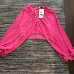 Rosa bolerotopp från Zara - Snygg rosa bolerotopp från Zara med långa ärmar och elastiska muddar. Perfekt för att lägga till en färgklick till din outfit. Toppen har en luftig design och är tillverkad i ett lätt material.