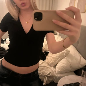 Svart v-ringad topp - Säljer en stilren svart v-ringad topp med korta ärmar. Toppen har en snygg omlott-design framtill som ger en elegant touch. Perfekt för att matcha med jeans eller kjol.