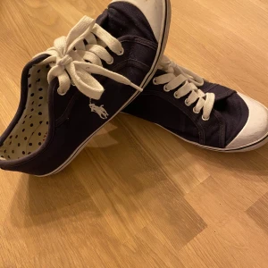 Mörklblå sneakers från Polo - Snygga mörk blå sneakers från Polo med vita snören och detaljer. Skorna har en klassisk design med en vit sula och prickigt mönster på insidan. Perfekta för en avslappnad stil.