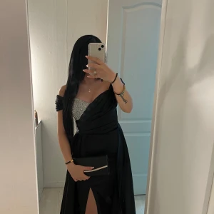 Svart långklänning med glittrig detalj - Elegant svart långklänning med en glittrig detalj på bröstet. Klänningen har en offshoulder-design och en slits framtill som ger en sofistikerad look. Perfekt för speciella tillfällen.