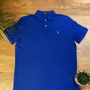 Blå pikétröja från Ralph Lauren - Säljer en klassisk blå pikétröja från Ralph Lauren i storlek M. Tröjan har en broderad orange logga på bröstet och är i mycket bra skick. Perfekt för både vardag och lite finare tillfällen. Skön och stilren design som aldrig går ur tiden!