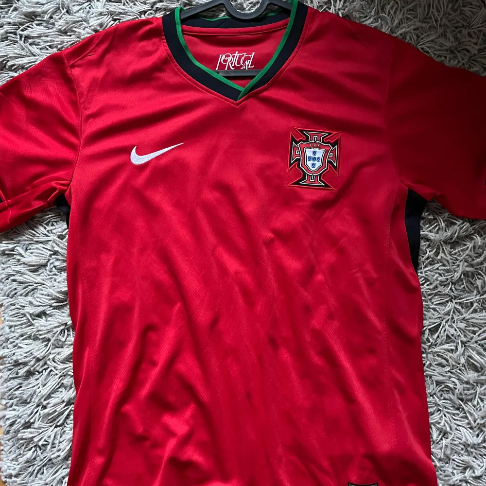 Säljer en röd Portugal fotbollströja från Nike i mycket bra skick. Tröjan har ett snyggt grönt och svart mönster runt halsen. . T-shirts.