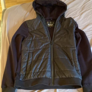 Svart jacka med huva från Barbour International - Säljer en snygg svart cardigan från Barbour International. Den har en quiltad framsida och mjuka ärmar, perfekt för höst och vår. Jackan har en dragkedja och en praktisk huva. Den är i mycket bra skick och passar perfekt för en casual look. Perfekt för dig som vill ha stil och komfort! Storleken är s och passar ungefär 175-185