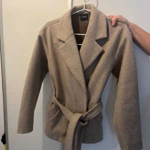 Beige kappa från vero moda!! - Säljer en stilren beige kappa från veromoda, använd ca 3 gånger. Den har en klassisk krage och ett bälte i midjan för en snygg passform. Perfekt för höst och vår! 🧥täcker precis rumpan, köpte för 800