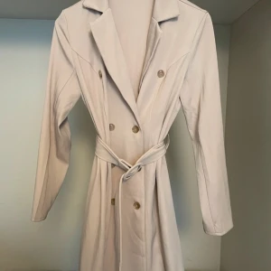 Beige trenchcoat med knappar - Säljer en stilren beige trenchcoat i mycket bra skick. Den har långa ärmar och ett bälte i midjan för en snygg passform. Perfekt för höst och vår! Knapparna framtill ger en klassisk look. Passar både till vardags och fest.