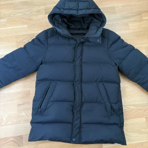 J.lindeberg vinterjacka  - Säljer en mörkblå/svart J.lindeberg pufferjacka med huva, perfekt för kalla dagar. Jackan har dragkedja och är långärmad. Den är i bra skick men har en liten fläck på ärmen som jag inte försökt få bort. Säljer jackan då den inte kommer till användning. Supermysig och varm för vintern!