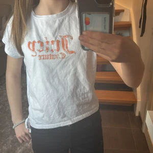 Vit t-shirt från Juicy Couture - Säljer en vit t-shirt från Juicy Couture med orange text på framsidan. Den har en fin snörning på sidan.T-shirten är i mycket bra skick och är i storlek xxs.Säljer för att den inte kommer till användning.