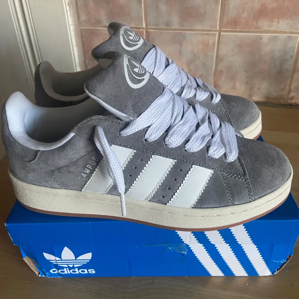 Säljer ett par snygga grå Adidas sneakers i mocka med vita detaljer och snörning. De har den klassiska tre-randiga designen på sidorna och Adidas-loggan på tungan och hälen. Perfekta för både vardag och fest! Skorna är i mycket bra skick och kommer i originalkartong. Sprillans nya och har inte kommit till användning 1 ända gång på grund av storleken. Skor.