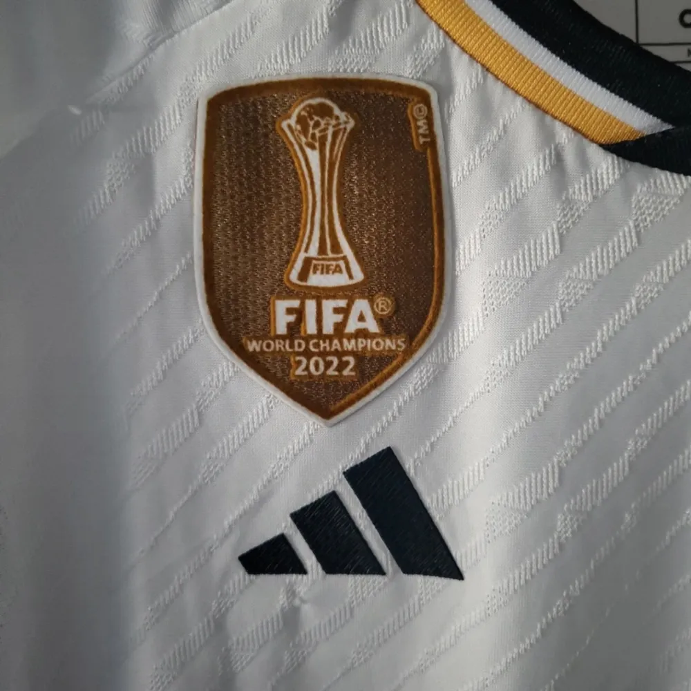 Säljer en vit Real Madrid fotbollströja i mycket bra skick. Tröjan har korta ärmar och är dekorerad med klubbens emblem och FIFA World Champions 2022-märke. Den har guldfärgade detaljer och är tillverkad av Adidas. Perfekt för alla fotbollsfans!. T-shirts.