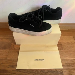 Skor från Axel Arigato - Säljer ett par snygga svarta sneakers från Axel Arigato i mycket bra skick. De har en stilren design med vit sula och är gjorda i mocka. Perfekta för både vardag och fest! Skorna kommer med originalförpackning.