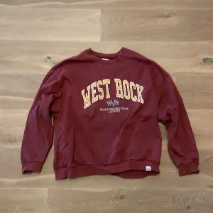 Säljer en vinröd sweatshirt med texten 'West Rock' i gult och vitt på framsidan. Tröjan är i bra skick och perfekt för en avslappnad stil. Den har långa ärmar och en bekväm passform. Perfekt för höst och vinter!