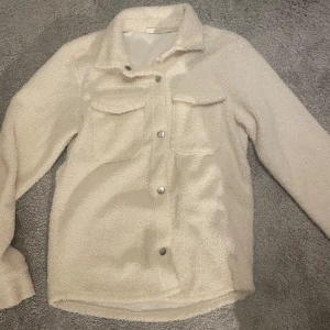 Vit teddyjacka från KappAhl - Mysig vit teddyjacka från KappAhl i storlek 146/152. Perfekt för kyliga dagar med sina långa ärmar och knappar framtill. Jackan har två praktiska bröstfickor och en skön krage. Passar både till vardags och lite finare tillfällen. Fint skick.