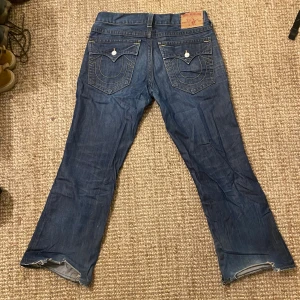 True Religion Billy - Säljer ett par blå jeans från True Religion i bra skick. De har en bootcut-stil och är tillverkade i USA. Jeansen har snygga bakfickor med knappar och slitna detaljer vid bensluten. Perfekta för en avslappnad look!