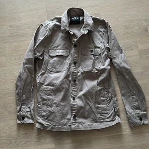 Beige overshirt/skjorta/jacka från Hollister - Säljer en snygg beige overshirt från Hollister i bra skick. Den har långa ärmar och knappar framtill samt på bröstfickorna. Strlk S