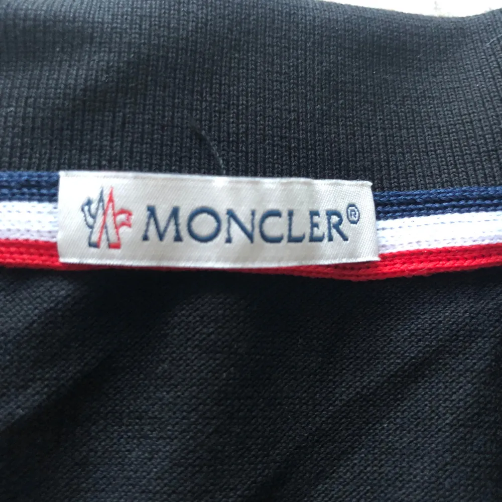 Säljer en snygg svart pikétröja från Moncler i nytt skick. Den har korta ärmar och en klassisk krage med röd och vit rand. Moncler-loggan är broderad på bröstet. Perfekt för en stilren och avslappnad look!. T-shirts.