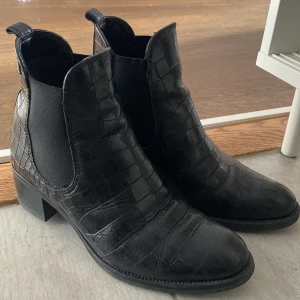 Svarta boots med krokodilmönster - Snygga svarta boots i skinn med krokodilmönster. De har en låg klack och elastiska paneler på sidorna för enkel på- och avtagning. Perfekta för både vardag och fest! 🖤