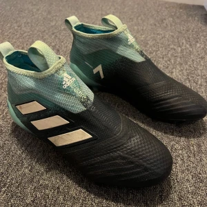 Adidas fotbollsskor i svart och mintgrön - Säljer ett par Adidas fotbollsskor i svart och mintgrön färg. Skorna har en modern design med de klassiska tre ränderna på sidan. De är i bra skick och perfekta för fotbollsträning eller match. Skorna är tillverkade i syntetmaterial och har en tight passform för optimal kontroll på planen.