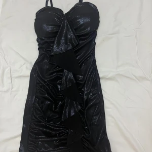 Svart glittrig klänning - Snygg svart glittrig klänning med axelband och en cool volangdetalj framtill. Perfekt för festliga tillfällen när du vill sticka ut. Klänningen har en tight passform som framhäver figuren.