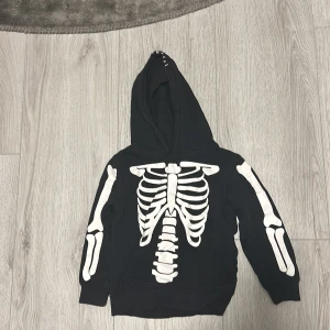 Svart hoodie med skelettmönster - Cool svart hoodie med ett vitt skelettmönster på framsidan. Perfekt för halloween. Den har en huva med ögon och tänder för en extra läskig look. Tröjan är i bra skick och passar perfekt för höst och vinter.