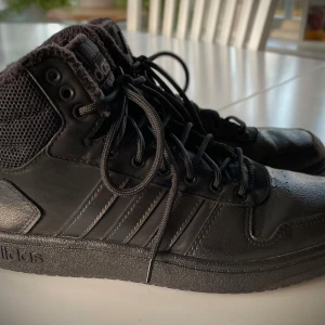 Svarta sneakers från Adidas - ADIDAS HOOPS 2.0 MID TRIPLE BLACK TUMBLED STRIPES  Säljer ett par svarta sneakers från Adidas i mycket bra skick. Snygg och stilren design med snörning och högre skaft. Mjukt, värmande innerfoder, passar bra i lite kallare temperaturer.  Sparsamt använda då sonen snabbt växte ur dem. Strl 42.                              Nypris 899 kr.          